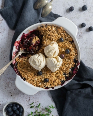 A large round dish of blueberry and thyme crumble with three scoops of vegan vanilla ice cream on top
