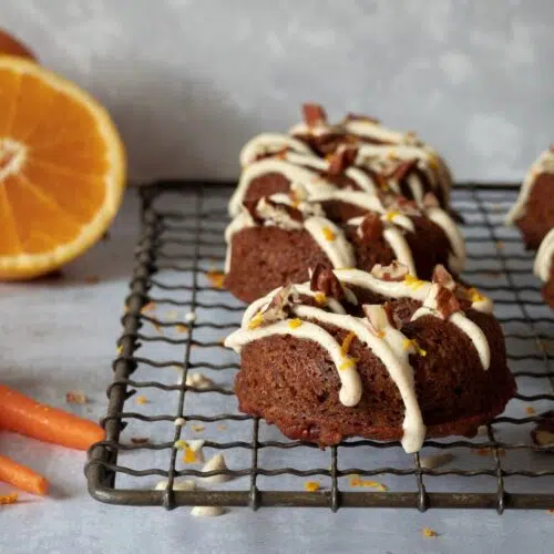 Baked carrot cake doughnuts lined up on a cooling rack with a zig zag of icing and crushed pecans on top