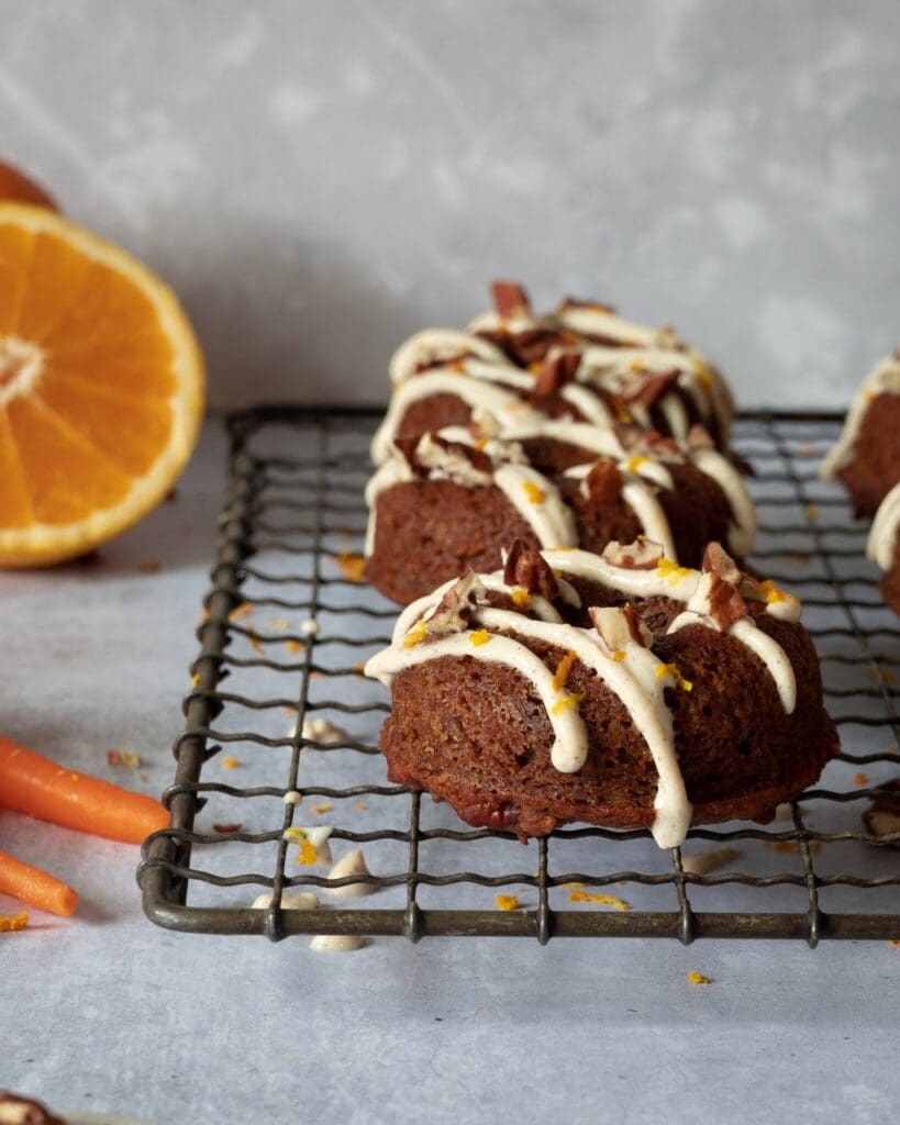 Baked carrot cake doughnuts lined up on a cooling rack with a zig zag of icing and crushed pecans on top