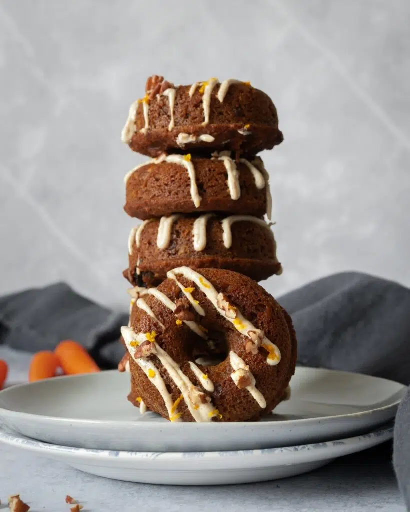 A stack of baked carrot cake doughnuts on a plate with a zig zag of icing over the top of each