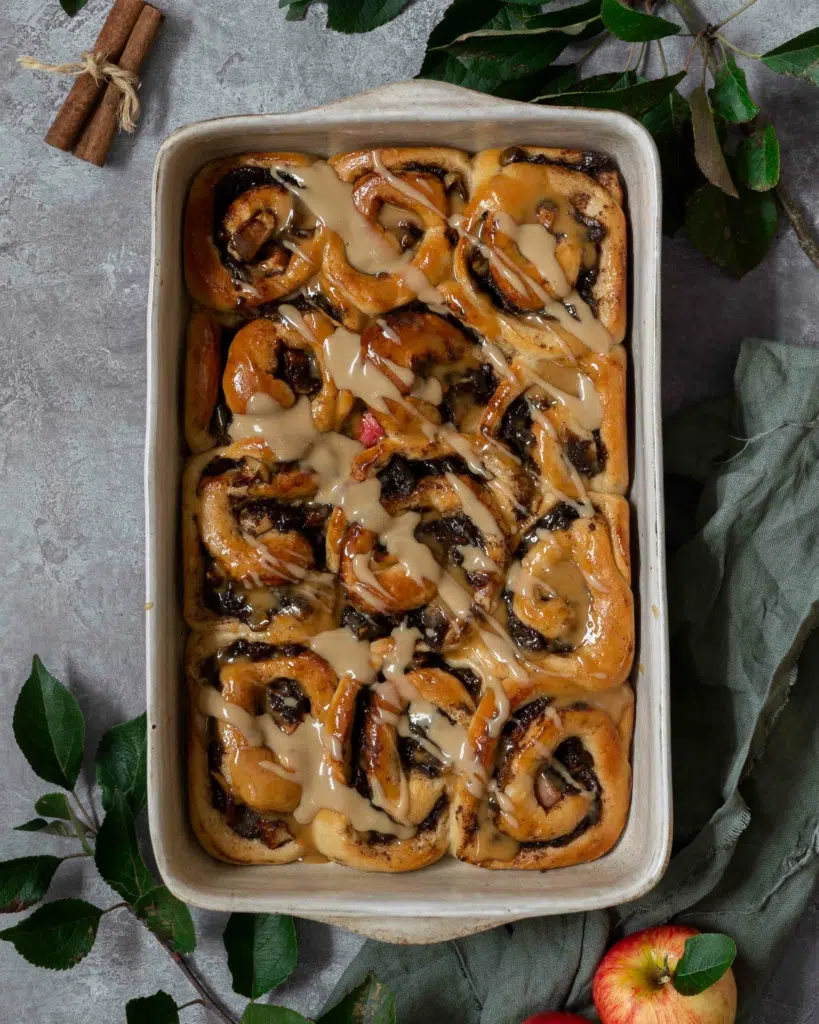 A tray of sticky, squishy cinnamon buns in a baking dish, studded with spiced cinnamon apple chunks and drizzled in creamy vegan caramel sauce