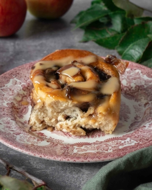 A sticky, squishy cinnamon bun sat on a pretty vintage pink plate, studded with spiced cinnamon apple chunks and drizzled in creamy vegan caramel sauce