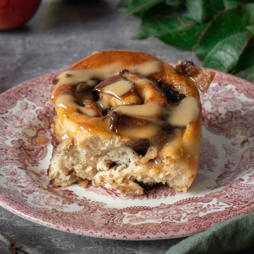 A sticky, squishy vegan apple cinnamon roll sat on a pretty vintage pink plate, studded with spiced cinnamon apple chunks and drizzled in creamy vegan caramel sauce