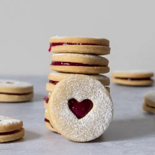 A stack of valentine's Jammie Biscuits on a table top dusted with icing sugar, with heart shaped cut outs displaying the raspberry jam in the centre.