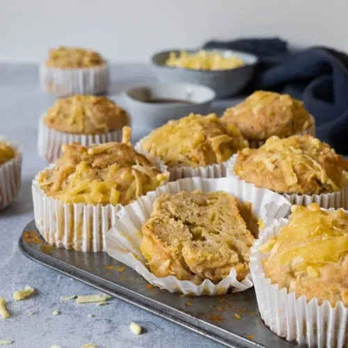A muffin tray filled with cheesy Welsh Rarebit Muffins, with one muffin cut in half displaying a squishy interior
