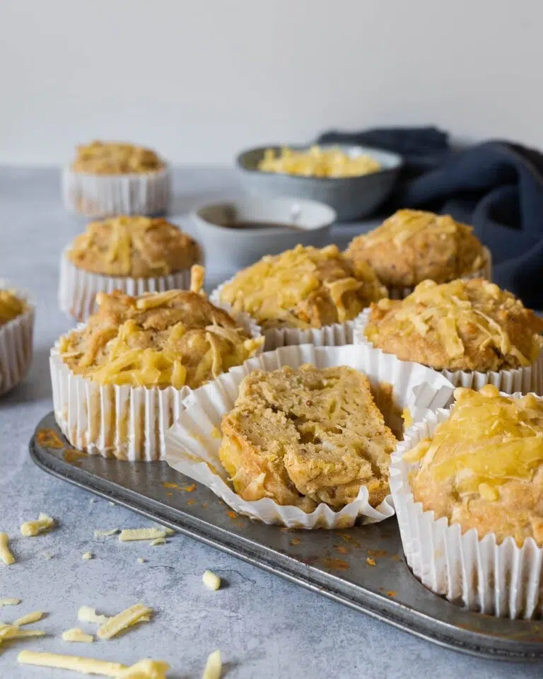 A muffin tray filled with cheesy Welsh Rarebit Muffins, with one muffin cut in half displaying a squishy interior