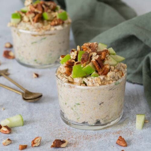 Two glass jars filled with creamy vegan bircher topped with crisp green apple, cinnamon, toasted pecans and maple syrup