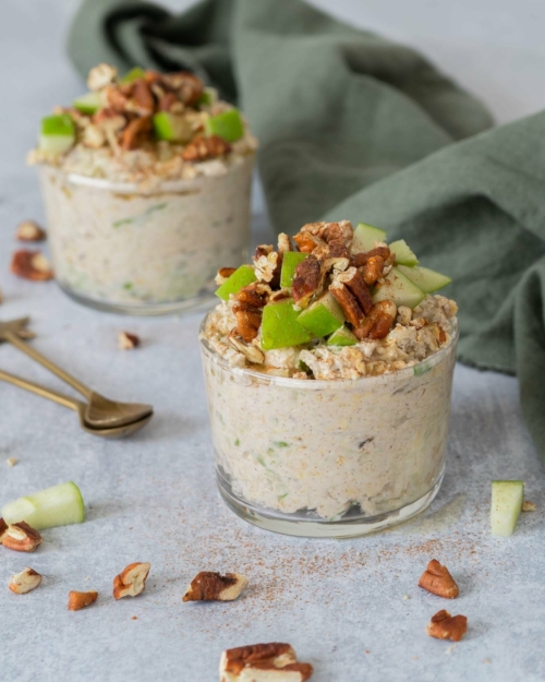 Two glass jars filled with creamy vegan bircher topped with crisp green apple, cinnamon, toasted pecans and maple syrup