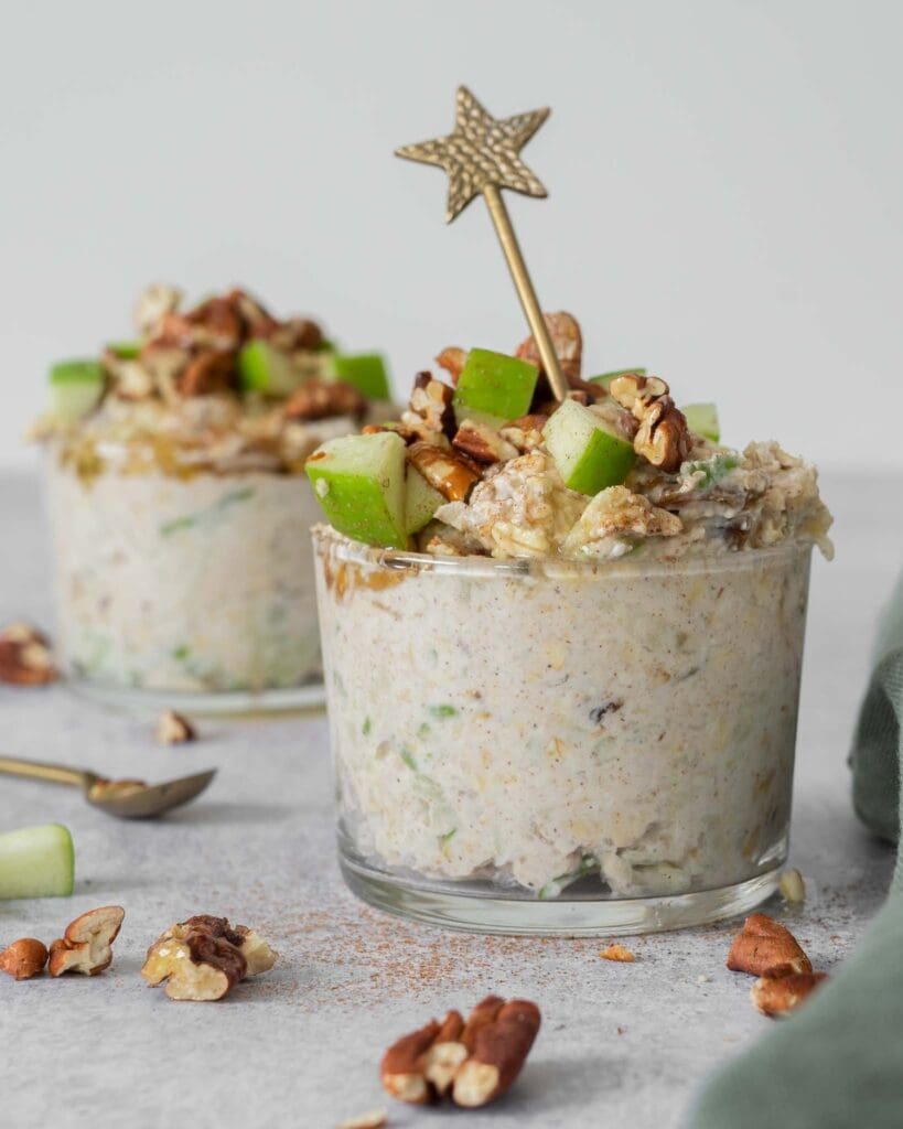 Two glass jars filled with creamy vegan bircher topped with crisp green apple, cinnamon, toasted pecans and maple syrup