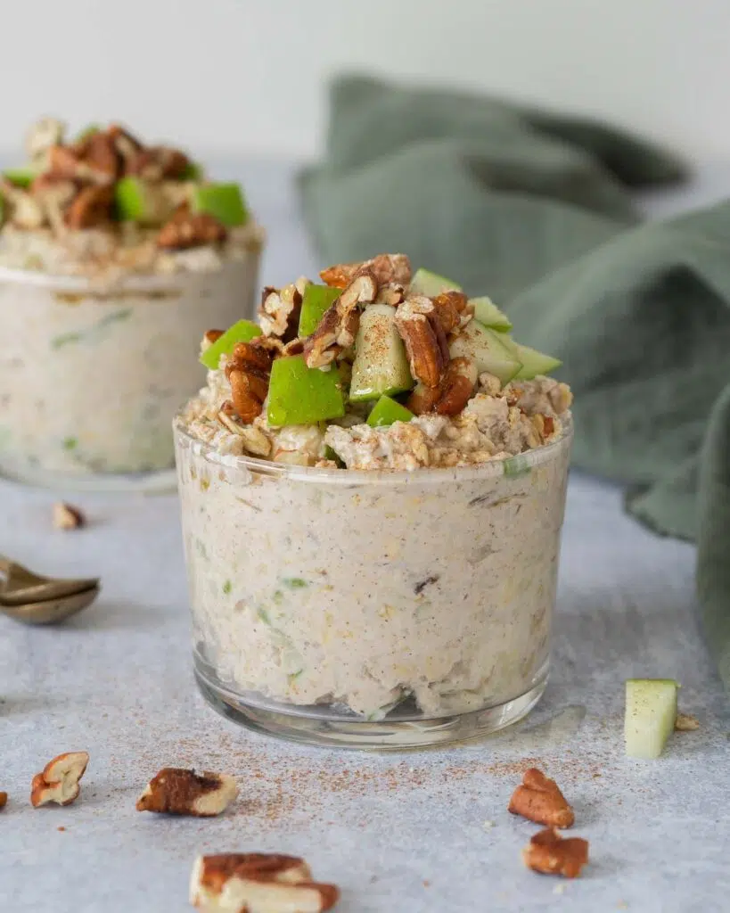 Two glass jars filled with creamy vegan bircher topped with crisp green apple, cinnamon, toasted pecans and maple syrup