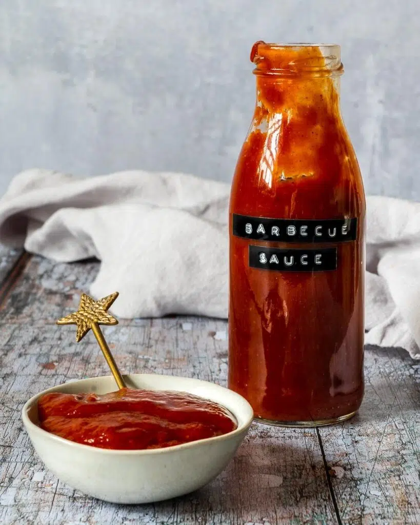 A glass bottle and small bowl filled with homemade rich red bbq sauce, with a gold star spoon sticking out of the bowl