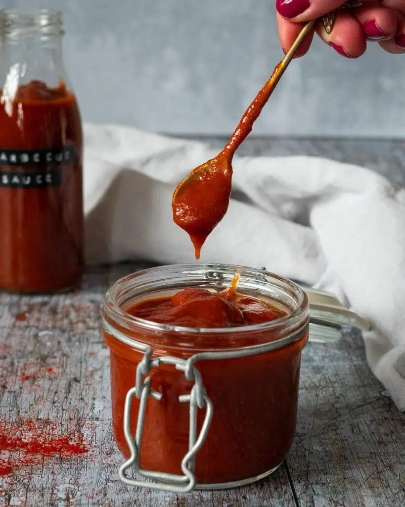 A glass jar filled with rich red bbq sauce, with a spoon lifting out of the jar