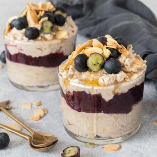 Two glass jars filled with creamy bircher, a blueberry puree layer and topped with fresh blueberries, flaked almonds and maple syrup