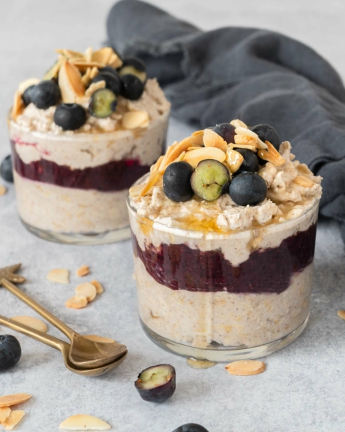 Two glass jars filled with creamy bircher, a blueberry puree layer and topped with fresh blueberries, flaked almonds and maple syrup