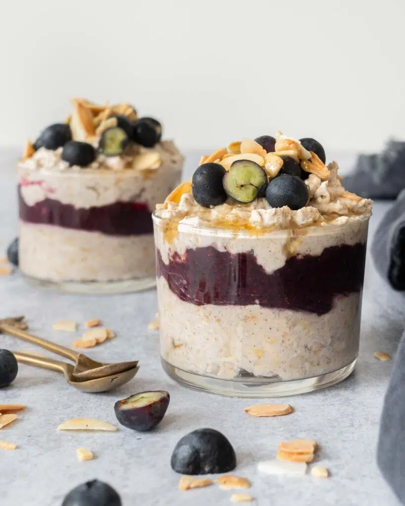 Two glass jars filled with creamy bircher, a blueberry puree layer and topped with fresh blueberries, flaked almonds and maple syrup