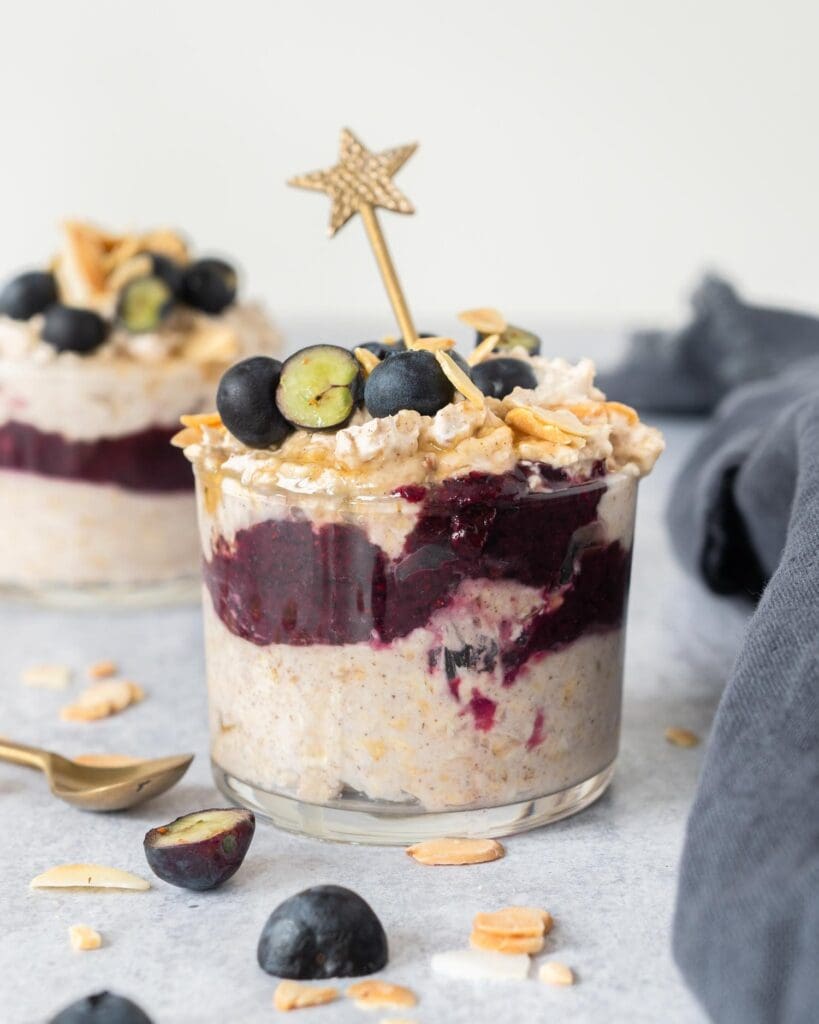 Two glass jars filled with creamy bircher, a blueberry puree layer and topped with fresh blueberries, flaked almonds and maple syrup