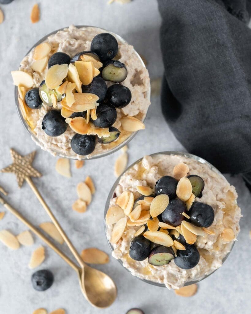 Two glass jars filled with creamy bircher, a blueberry puree layer and topped with fresh blueberries, flaked almonds and maple syrup