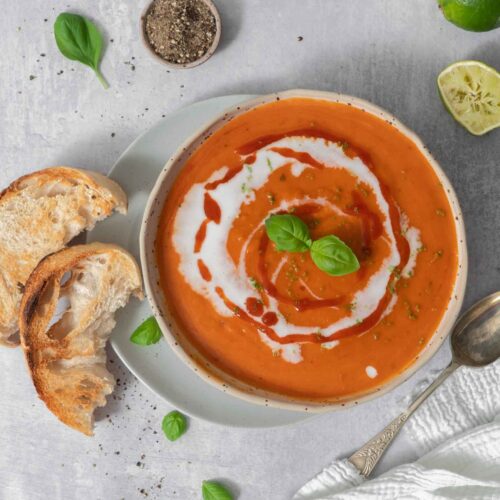 A top down look at a bowl of warming, rich, butternut squash and roasted red pepper soup in a rich orange colour, drizzled with vegan cream, sriracha and topped with fresh basil leaves. Served with a side of sourdough toast.