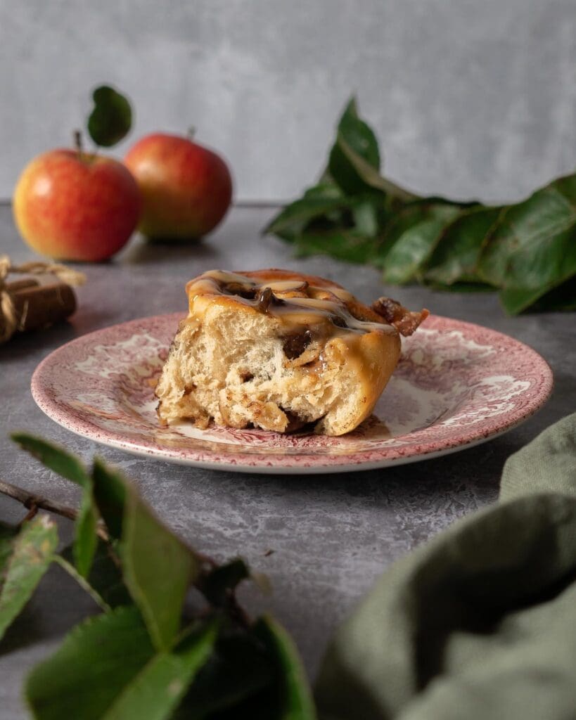A sticky caramel apple cinnamon bun sat on a vintage pink plate, surrounded by freshly picked apples, apple tree leaves and cinnamon sticks.