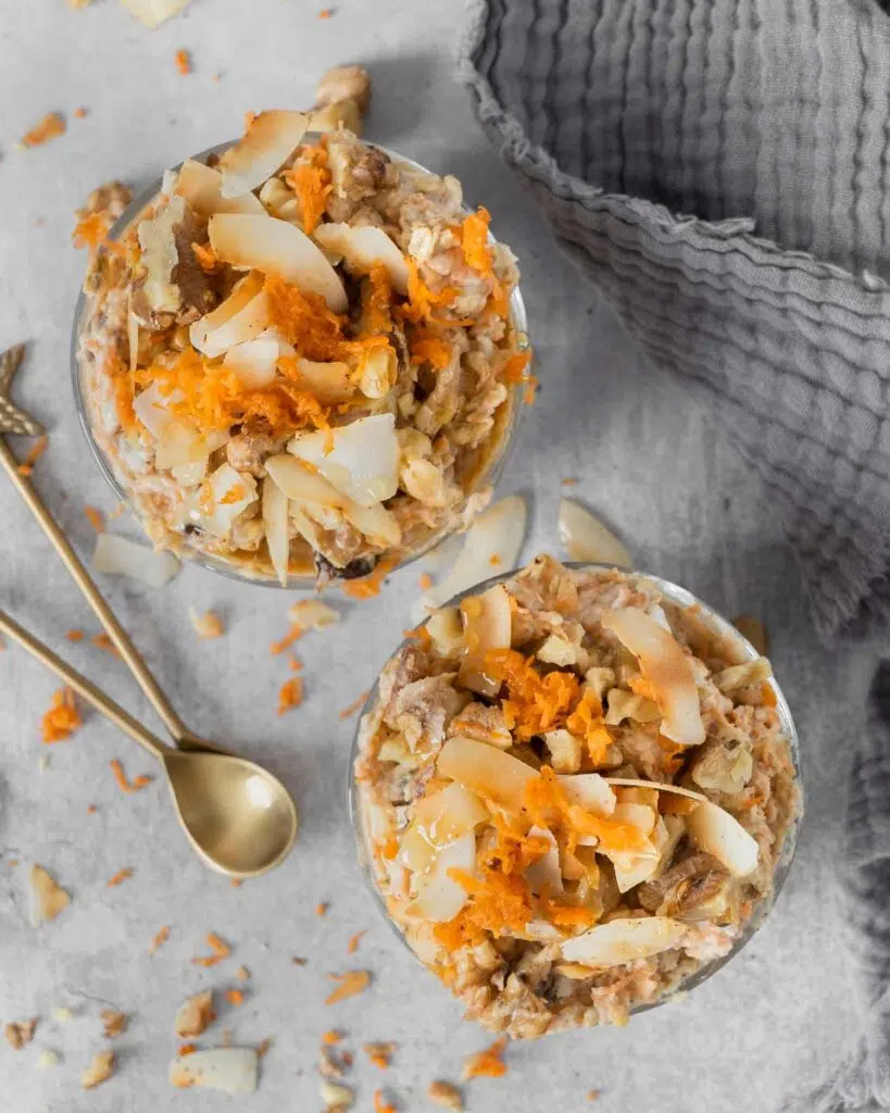 Two glass jars filled with creamy vegan bircher topped with grated carrot, walnuts and toasted coconut flakes