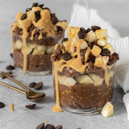 Two glass dishes overflowing with creamy chocolate peanut bircher topped with fresh banana, chocolate chips and a peanut butter drizzle
