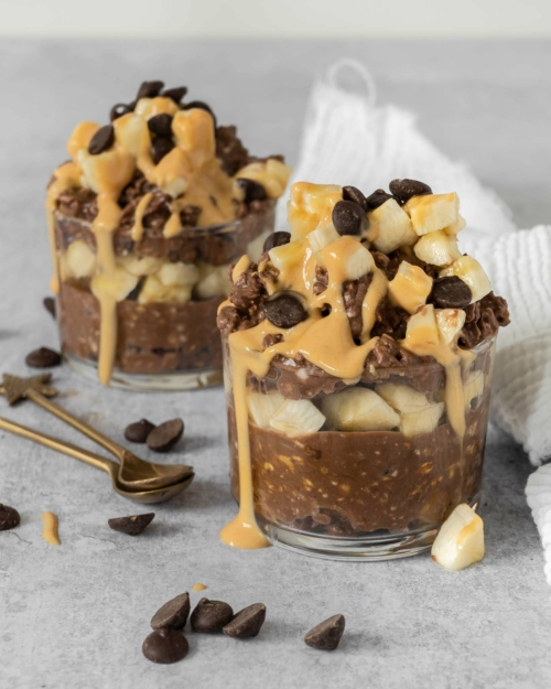 Two glass dishes overflowing with creamy chocolate peanut bircher topped with fresh banana, chocolate chips and a peanut butter drizzle