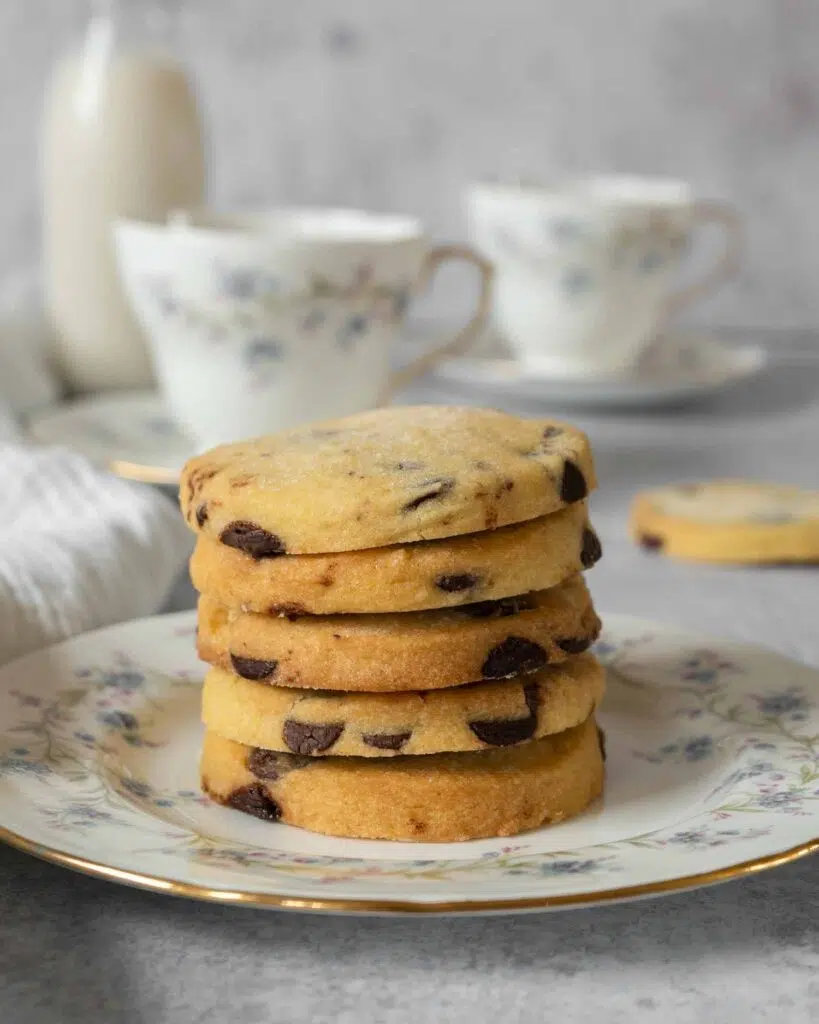 A stack of buttery shortbread cookies studded with dark chocolate chips and dusted in sugar, resting on a pretty floral vintage tea plate