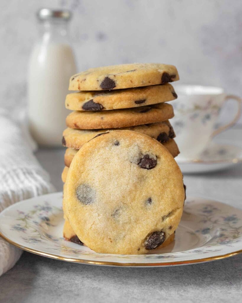 A stack of buttery shortbread cookies studded with dark chocolate chips and dusted in sugar, resting on a pretty floral vintage tea plate