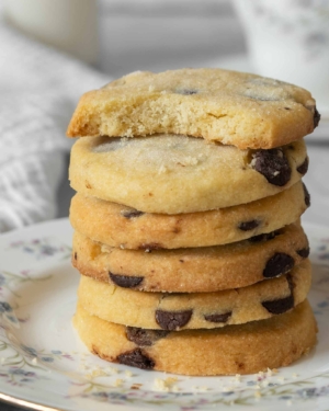 A stack of buttery shortbread cookies studded with dark chocolate chips and dusted in sugar, resting on a pretty floral vintage tea plate. The top cookie has a bite taken out of it, displaying a crumbly, buttery interior.