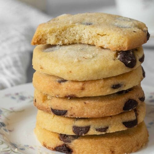 A stack of golden chocolate chip shortbread cookies sprinkled in sugar. The top cookie has had a bite taken out of it, revealing the crumbly, buttery texture inside.