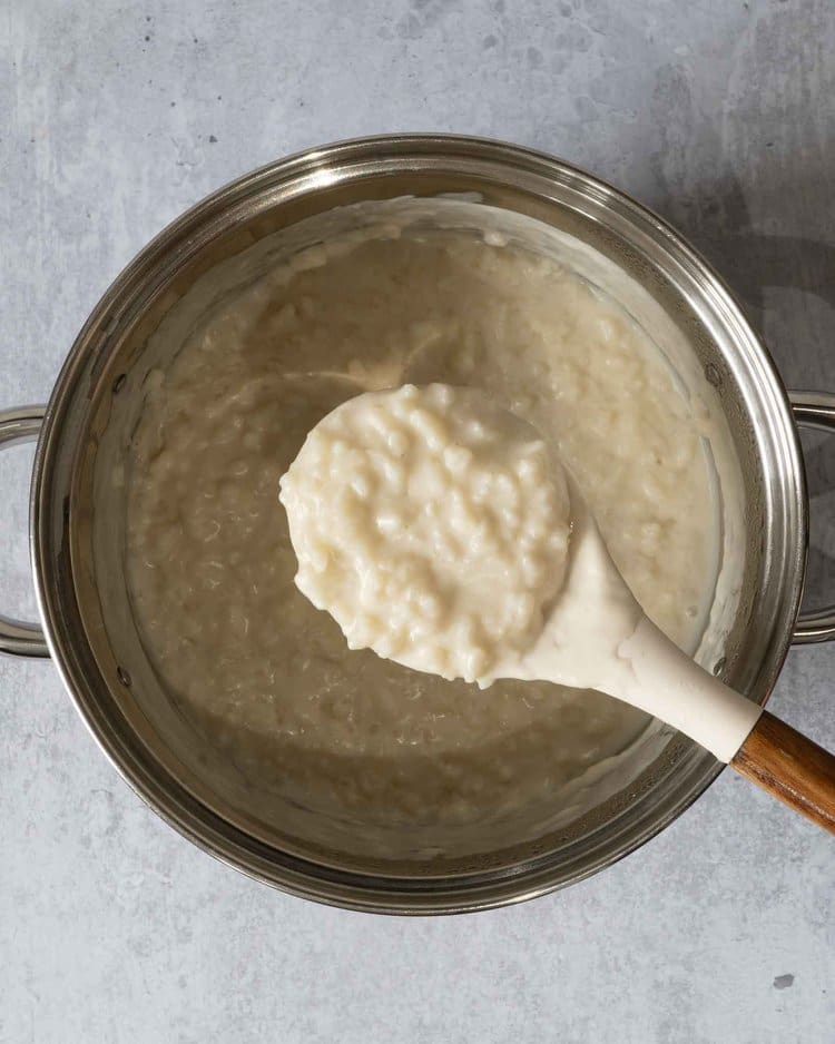 The ingredients for making coconut rice pudding in a saucepan with a spoonful of creamy rice pudding being scooped out