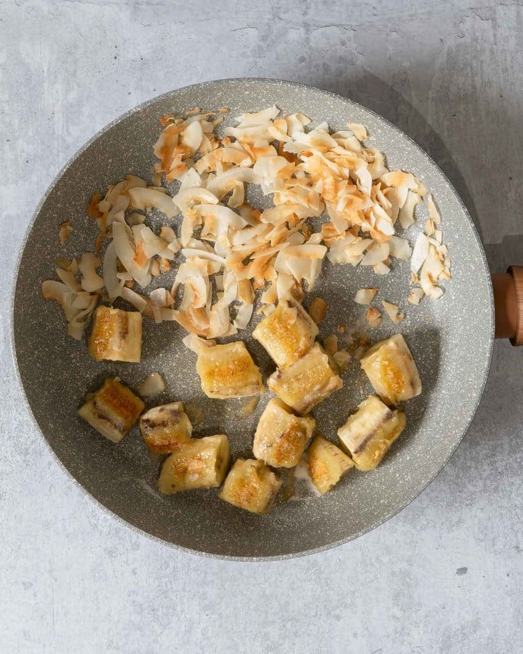 A non stick frying pan filled with toasted golden coconut flakes and sticky caramelised banana chunks