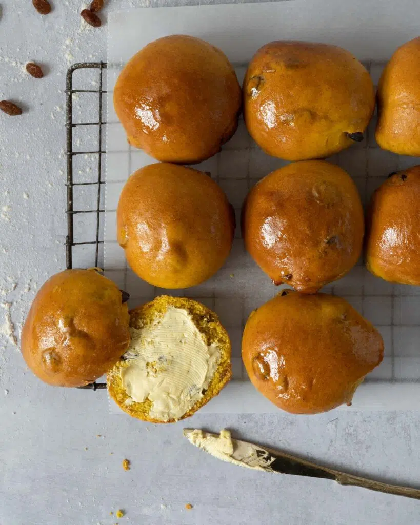 Glistening golden saffron buns cooling on a rack, one cut open with vegan butter slathered on.