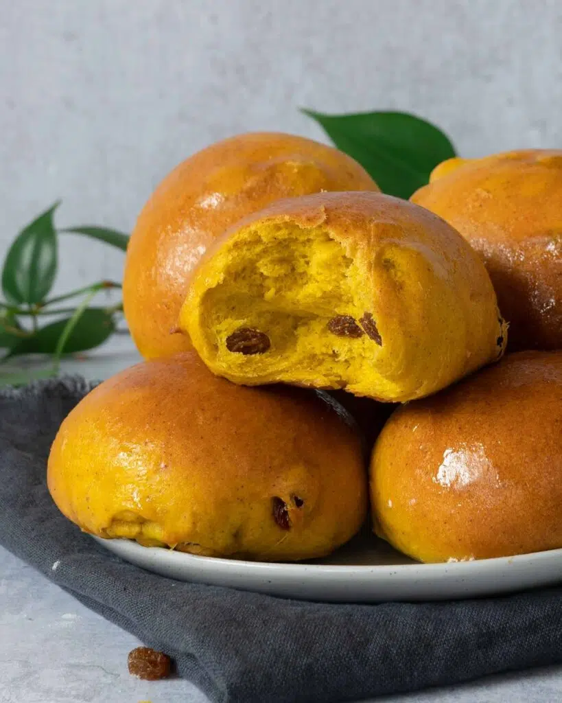 Glistening golden Cornish saffron buns stacked on a plate, one with a bite taken out displaying the inside studded with juicy sultanas