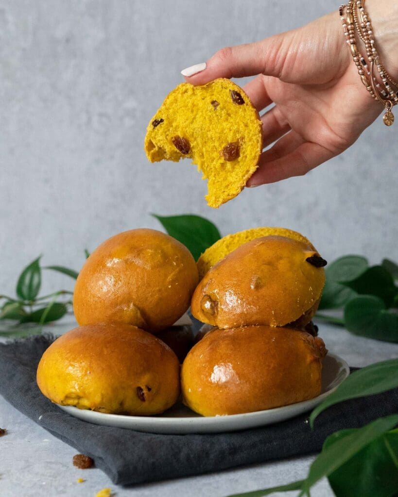 Glistening golden Cornish saffron buns stacked on a plate, the top one cut in half displaying a soft interior studded with juicy sultanas