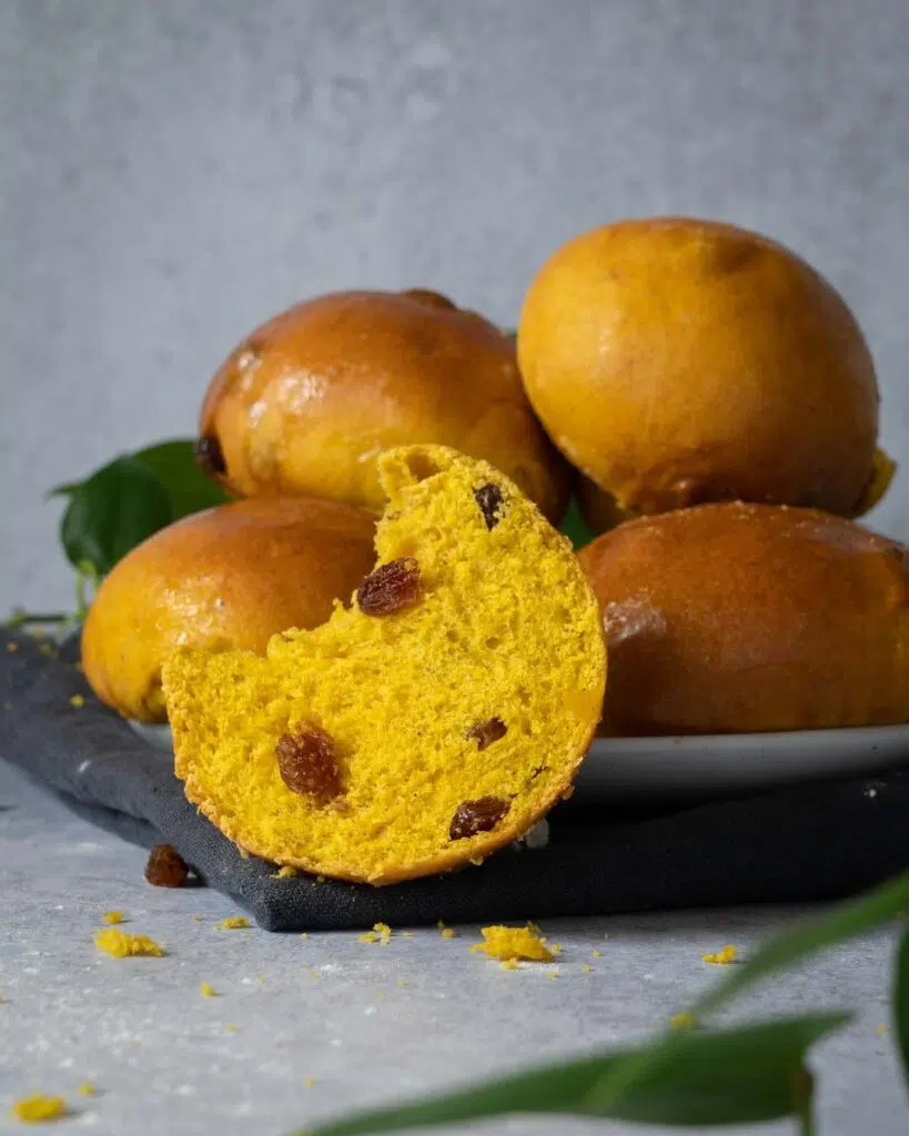 Glistening golden Cornish saffron buns stacked on a plate, one cut in half displaying a soft interior studded with juicy sultanas