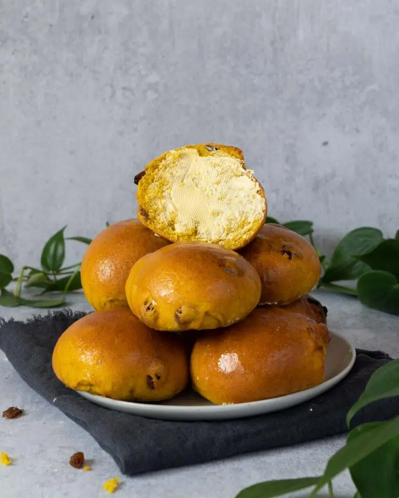 Glistening golden Cornish saffron buns stacked on a plate, the top one cut in half and slathered with vegan butter
