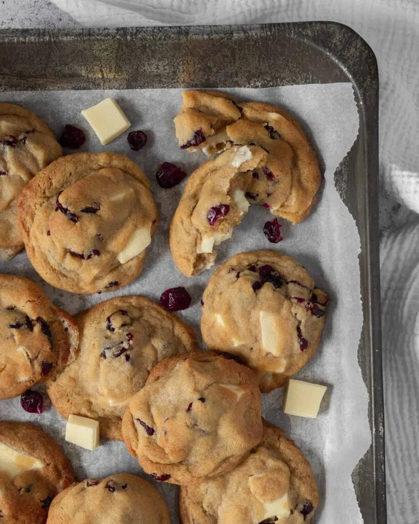 Cranberry and white chocolate cookies in a baking tin surrounded by chunks of vegan white chocolate and juicy, tangy red cranberries