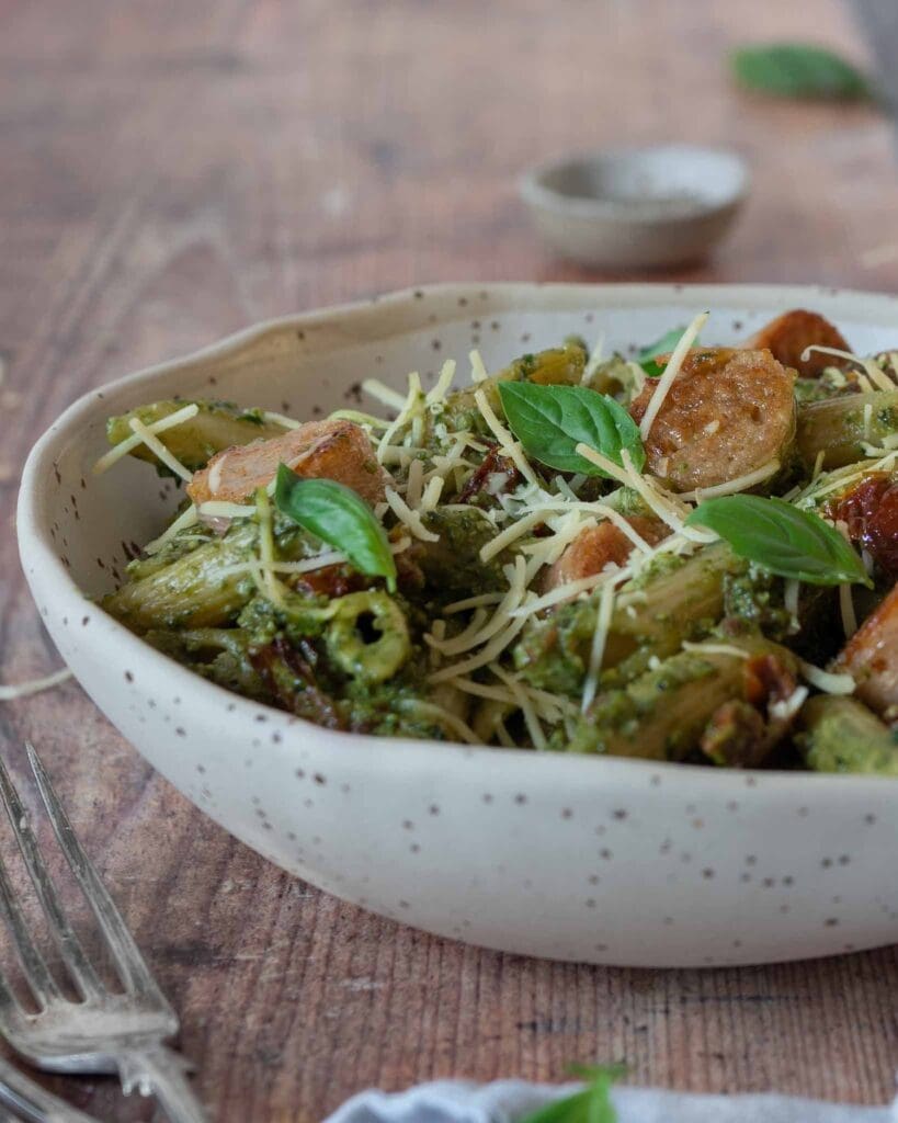 Pesto pasta with sun-dried tomato pieces stirred through, topped with crispy vegan sausage slices, dairy free cheese and fresh basil leaves