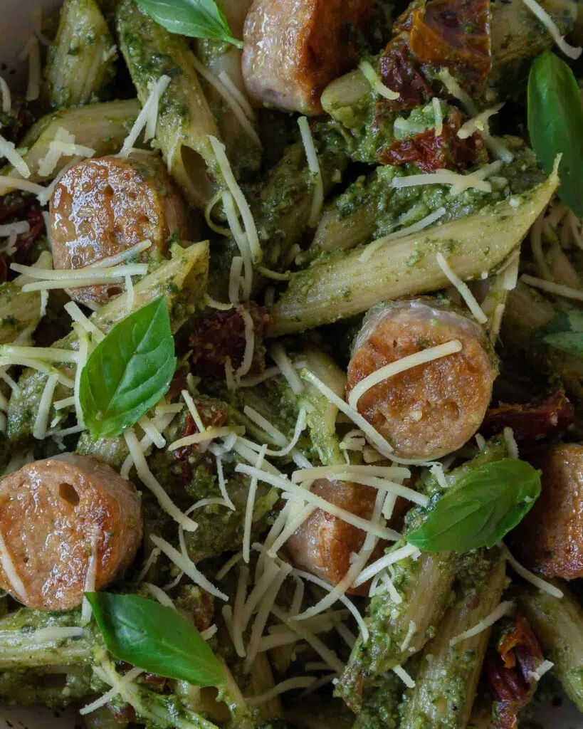 Pesto pasta with sun-dried tomato pieces stirred through, topped with crispy vegan sausage slices, dairy free cheese and fresh basil leaves