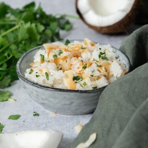 A bowl of fluffy white coconut rice topped with lightly toasted coconut flakes and fresh coriander. Fresh coconut and a bunch of fresh coriander surround the bowl.
