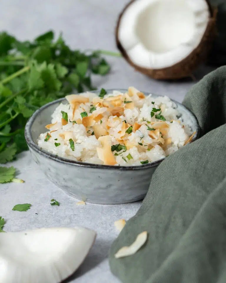 A bowl of fluffy white coconut rice topped with lightly toasted coconut flakes and fresh coriander. Fresh coconut and a bunch of fresh coriander surround the bowl.
