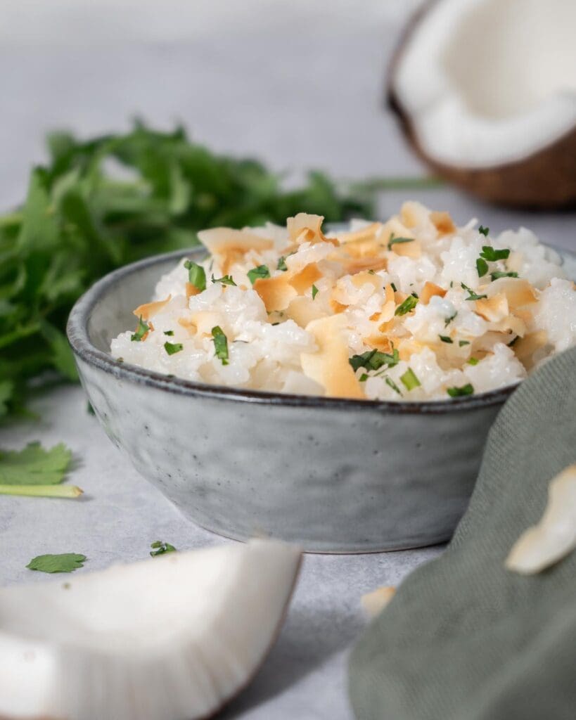 A bowl of fluffy white coconut rice topped with lightly toasted coconut flakes and fresh coriander.  Fresh coconut and a bunch of fresh coriander surround the bowl.