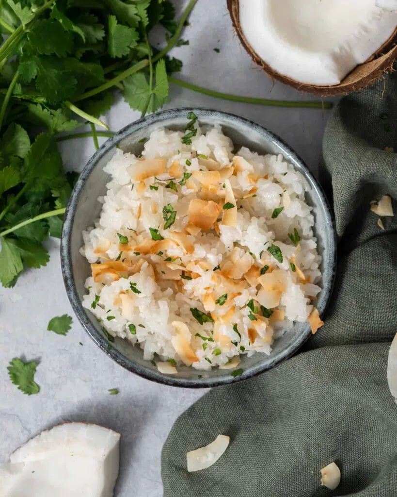 A bowl of fluffy white coconut rice topped with lightly toasted coconut flakes and fresh coriander.  Fresh coconut and a bunch of fresh coriander surround the bowl.