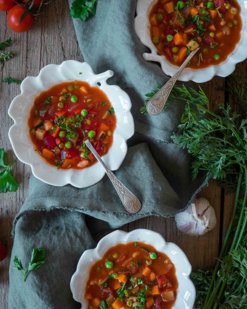 Three bowls of colourful farmhouse vegetable soup on a rustic wooden farmhouse table with fresh herbs, carrots, tomatoes and garlic scattered around