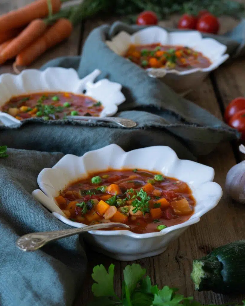 Three bowls of colourful farmhouse vegetable soup on a rustic wooden farmhouse table with fresh herbs, carrots, tomatoes and garlic scattered around