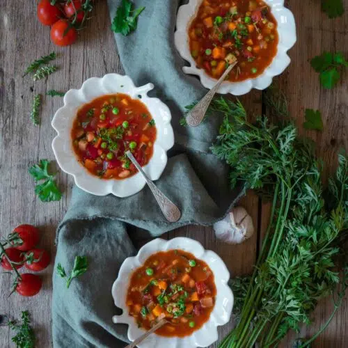 Three bowls of colourful farmhouse vegetable soup on a rustic wooden farmhouse table with fresh herbs, carrots, tomatoes and garlic scattered around