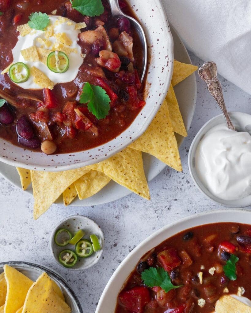 Two bowls of jackfruit and three bean chilli soup, in a rich deep red colour with pops of green coriander, topped with vegan creme fraiche  and crumbled tortilla chips