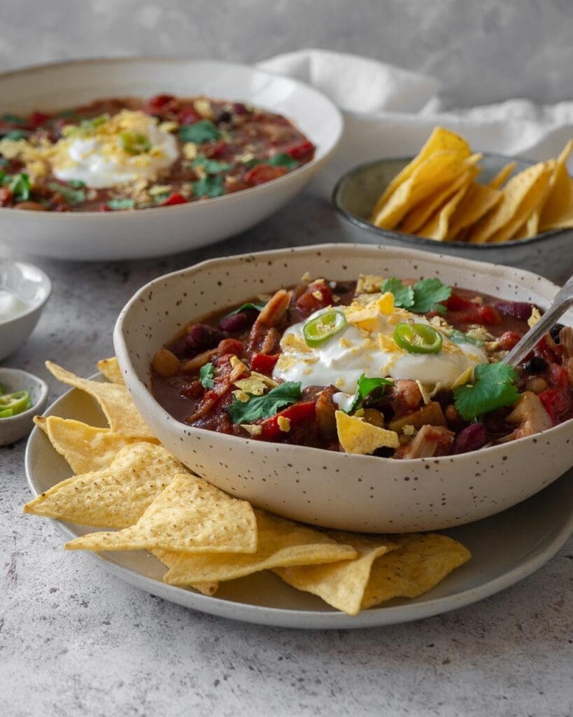 Two bowls of jackfruit and three bean chilli soup, in a rich deep red colour with pops of green coriander, topped with vegan creme fraiche  and crumbled tortilla chips