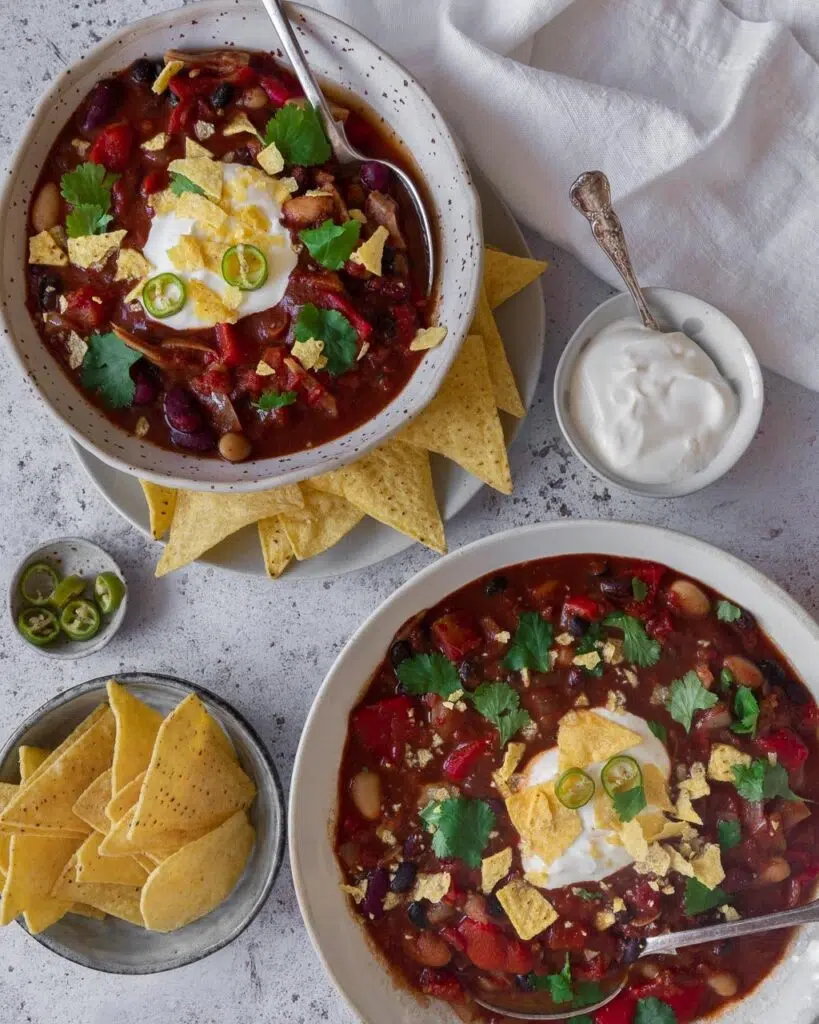 Two bowls of jackfruit and three bean chilli soup, in a rich deep red colour with pops of green coriander, topped with vegan creme fraiche  and crumbled tortilla chips